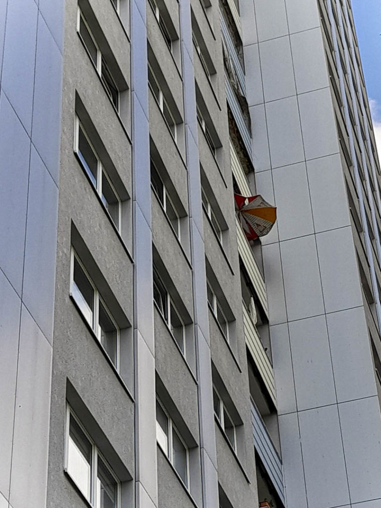 Foto eines zotteligen Sonnenschirms, der aus einem Fenster im Salvador-Allende-Plattenbau ragt