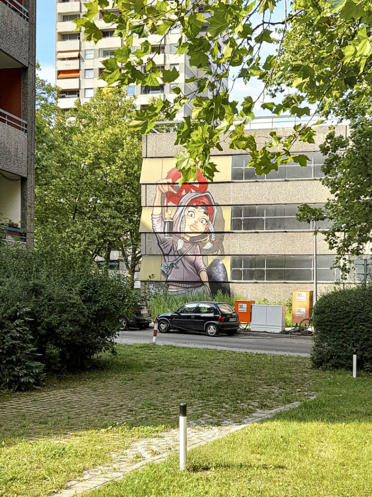 Foto eines Grafitto mit einem coolen Mädchen, das eine Spraydose fallenlässt, in der Gropiusstadt, drumherum grüne Bäume und Hochhäuser